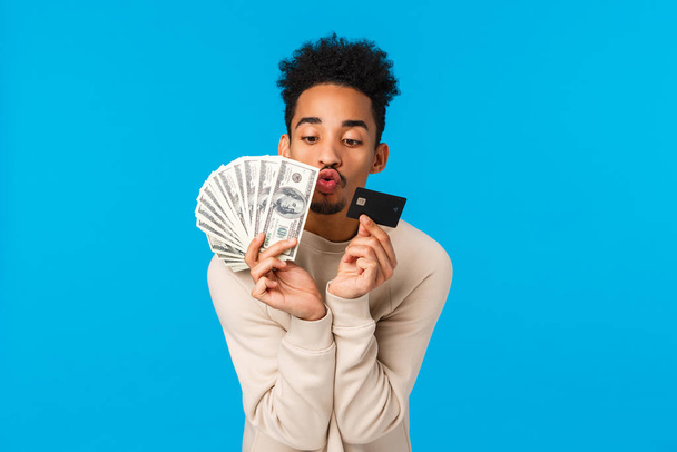 Waist-up portrait addicated and pleased african-american guy looking at precious money and credit card, like waisting cash on winter holidays sale season, kissing them, blue background - Photo, Image