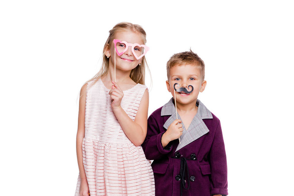 Dívka s papírovými brýlemi ve tvaru srdce a chlapec s papírovým knírkem pózující na bílém pozadí - Fotografie, Obrázek