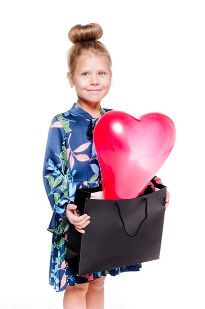 Little fashionable girl in a blue floral dress holds a large bag with a heart-shaped balloon inside - Zdjęcie, obraz