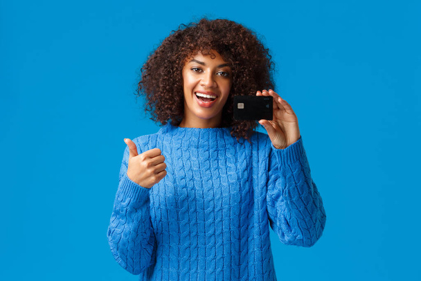 Satisfied young modern girl got bank account, opened deposit, using cashback service. Attractive african american woman in winter sweater, show thumbs-up and credit card in approval, recommend - Photo, Image