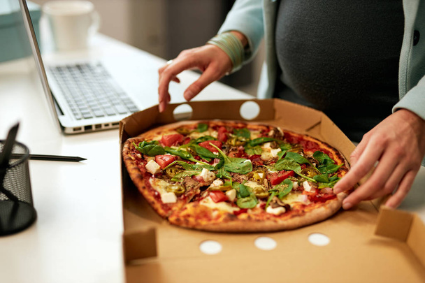 Hladová běloška těhotná podnikatelka stojící v kanceláři a pije pizzu k obědu. - Fotografie, Obrázek