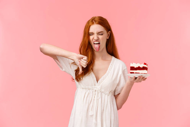 Tudatlan, válogatós és sznob vörös hajú szülinapos lány utálja a b-napos tortát, tányért tart desszerttel, nyelves grimaszolást mutat, és hüvelykujját lefelé a rosszallás, idegenkedés és undor - Fotó, kép