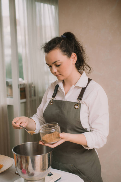 A cozinheira derrama açúcar de cana marrom em uma tigela de metal. Cozinhar doces na cozinha
 - Foto, Imagem