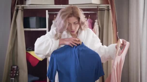 Portrait of young confident girl trying on blue shirt and pink blouse. Beautiful woman in white bathrobe choosing elegant outfit in the morning. Style, fashion. - Séquence, vidéo
