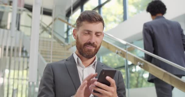 Close up of bearded businessman using smartphone and standing in modern office. Male company chief surfing net and swiping screen his mobile phone. Concept of people, work, technology. - 映像、動画