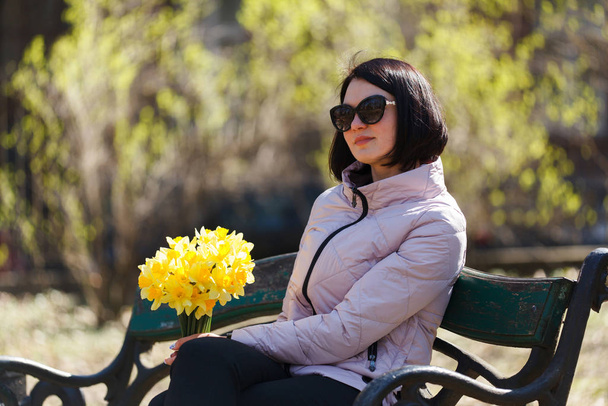 Güneşten gözlük güzel kız bankta parkta oturan nergis bir demet tutar. Bir avuç çiçek - Fotoğraf, Görsel