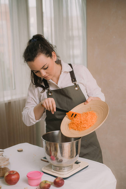 O cozinheiro adiciona as cenouras raladas à tigela de metal. Mão feminina colocar cenoura picada em tigela de madeira com salada na cozinha. Cozinhar legumes
 - Foto, Imagem