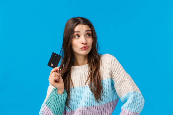 Shopping, winter holidays and women concept. Curious and thoughtful good-looking stylish woman thinking how waist all money, holding credit card, pouting and pondering, blue background - 写真・画像