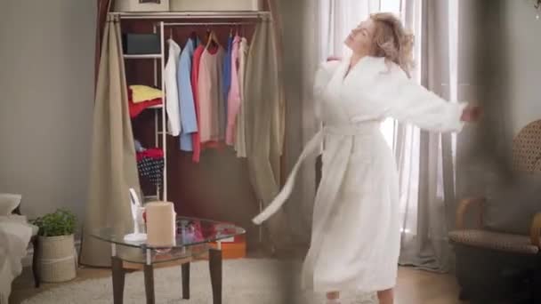 Young Caucasian woman in white bathrobe dancing and gesturing at home. Happy female melomaniac enjoying music on weekends. Hobby, lifestyle, happiness. - Filmagem, Vídeo