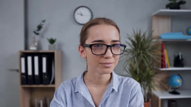 Front view of good-tempered smiling blond woman in glasses which posing on camera ,sitting in modern room - Materiaali, video