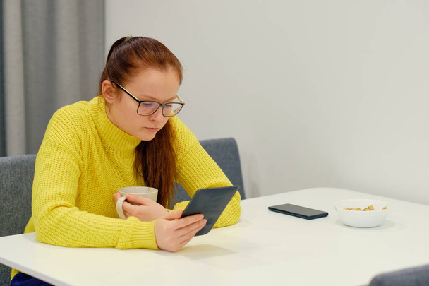 Молода біла жінка в жовтих в'язаних светрних сидіннях за столом в сучасному світлому інтер'єрі і читає електронну книгу. Концепція освіти, підготовка до іспиту, читання, кава, сніданок
 - Фото, зображення