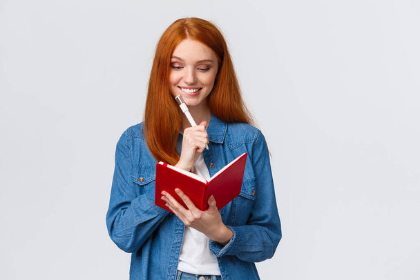Creative smiling cute redhead woman, had romantic date, writing it down in secret diary, grinning and blushing, look pleased pages of notebook, standing white background - Fotó, kép