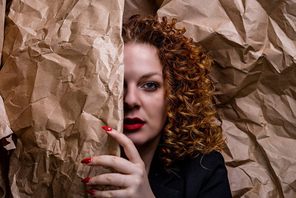 Mooi meisje met krullend rood haar met rode lippen, bedekt met een verfrommeld vel kraftpapier voor verpakking op de achtergrond van verfrommeld kraftpapier - Foto, afbeelding