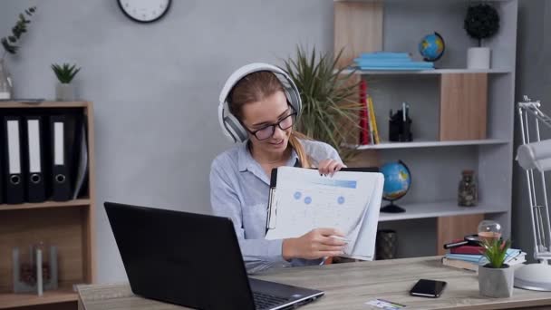 Close view of splendid blond 25-aged woman in headphones with financial reports in hands which having videocall with client - Filmati, video