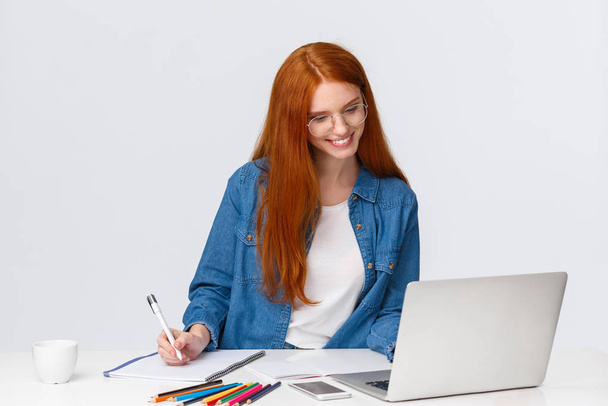 Education, work and vanlife concept. Cheerful lovely redhead woman in glasses standing near table, reading something from laptop, gather information from internet to write good essay - Photo, Image