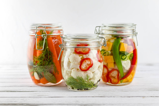 drie glazen potjes gefermenteerde bloemkool, wortelen, chili peper, groenten op een lichte ondergrond. fermentatie is een bron van probiotica - Foto, afbeelding