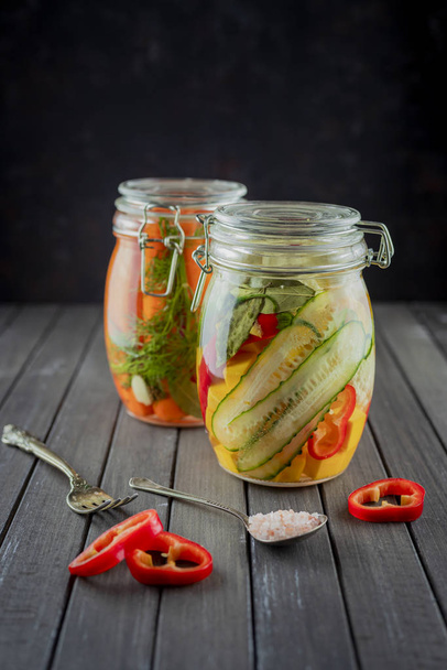 two glass jars of fermented cucumbers, carrots, pepper, salt, vegetables on a dark background.fermentation is a source of probiotic. popular vegetarian food. copy space - Foto, Imagen