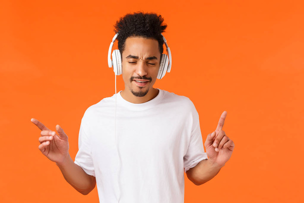 Man enjoying awesome beats. Attractive modern hipster african-american guy with afro haircut, moustache, close eyes drumming with fingers and listen music in headphones, orange background - Fotoğraf, Görsel