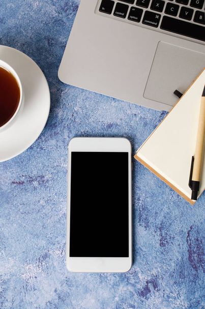 Λευκό smartphone με μαύρη κενή οθόνη στο γραφείο με φορητό υπολογιστή, άδειο σημειωματάριο και φλιτζάνι τσάι. Mock up του τηλεφώνου. - Φωτογραφία, εικόνα
