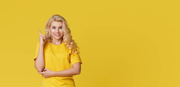 Zdjęcie szczęśliwej młodej blondynki stojącej samotnie nad żółtą ścianą. Patrząc na kamerę pokazującą kierunek miejsca kopiowania. Sztandar - Zdjęcie, obraz