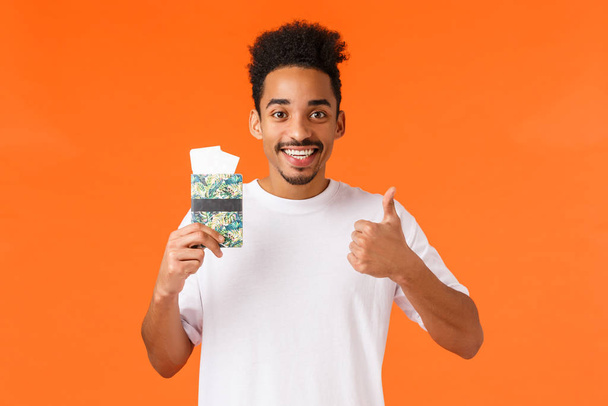 Удовлетворенный хорошо выглядящий афроамериканец-турист, улыбающийся и дающий разрешение, рекомендующий турагентство, имеющий паспорт с двумя билетами, стоящий оранжевый фон
 - Фото, изображение