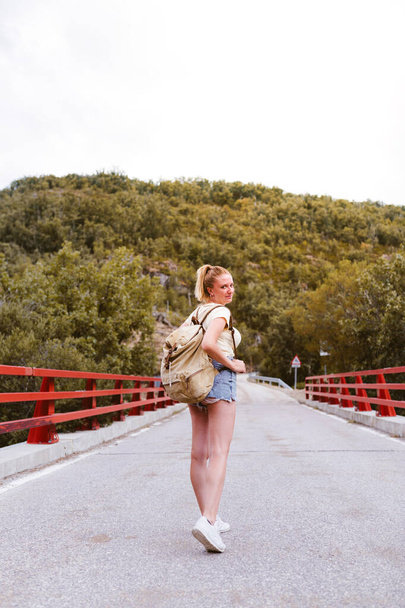 Back view of young blonde woman with backpack walking on the road over a bridge near the mountain. Travel and adventure concept. Traveler in the middle of woodland. Travel alone - Foto, immagini