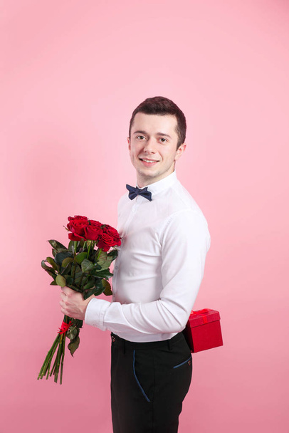Young handsome man with beautiful flower bouquet and red gift box on pink background - Foto, afbeelding
