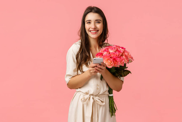 Technology, romance and happiness concept. Tender young cheerful smiling woman with beautiful flowers, holding smartphone, answer on congratulations b-day, chatting on birthday, pink background - Photo, Image