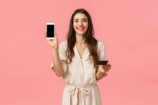 Vyötärö-up muotokuva onnellinen typerä nauraa eurooppalainen nainen Varauksen verkossa, osoittaa viileä ostoksia sovellus älypuhelimen näytöllä, tilalla matkapuhelin ja luottokortti, vaaleanpunainen tausta - Valokuva, kuva