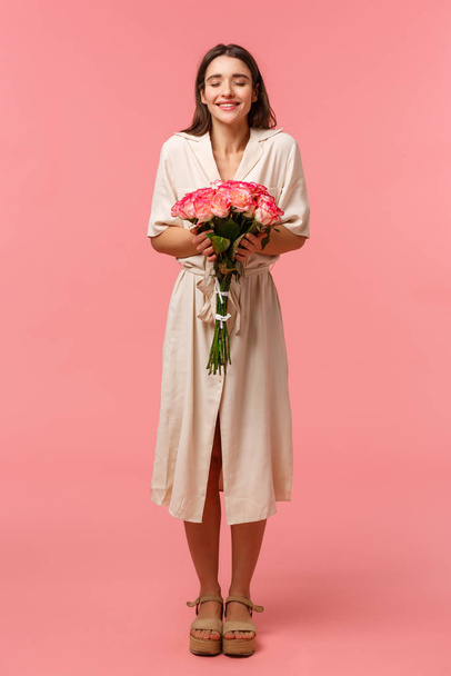 Full-length κάθετο πορτρέτο της ονειρικής, ρομαντική γυναίκα στο φόρεμα, λαμβάνοντας όμορφα τριαντάφυλλα από μυστικό θαυμαστή για τα γενέθλια, κλείστε τα μάτια και οσμή των λουλουδιών, στέκεται ροζ φόντο - Φωτογραφία, εικόνα