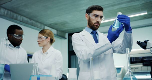 Caucasian handsome medical laboratory male worker doing analysis of some liquid in the test tubes while his multiethnical co-workers talking on the background. - 写真・画像