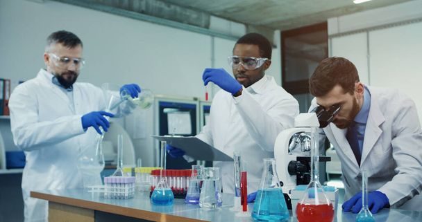 Portrait shot of the  men scientist looking in the microscope while investigating something in the laboratory, while his male mixed-races co-workers talking on the background. - Фото, изображение