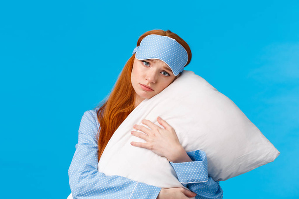 Sad redhead girl feeling depressed dont want college, lying in bed, hugging pillow, frowning and looking uneasy with concerned, nervous expression, wearing sleep mask and nightwear, blue background - Photo, Image