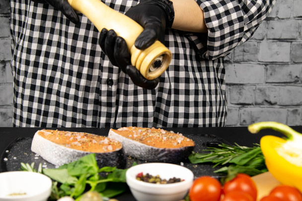 Les mains préparent et salent le steak de saumon pour la cuisson, à côté du basilic de table, des épices au romarin et des poivrons
 - Photo, image