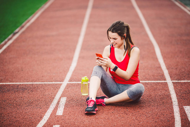 beautiful fitness girl uses smartphone at stadium after workout. Sports and healthy. Sport woman use of cellphone inside sport complex. Sport and technology. resting after workout, run at red track - Zdjęcie, obraz