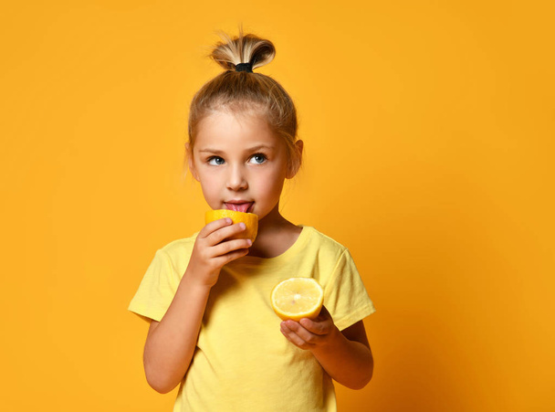 Little cute blond girl in yellow t-shirt holding and licking half of fresh ripe lemon fruit over yellow background - Foto, afbeelding