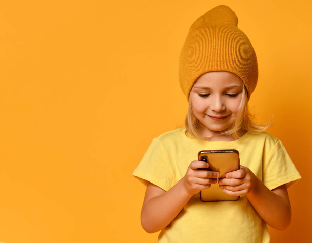 Little blonde kid dressed in t-shirt and hat, posing with smartphone against yellow background. Technology, children, internet. Close-up shot - Foto, Imagem