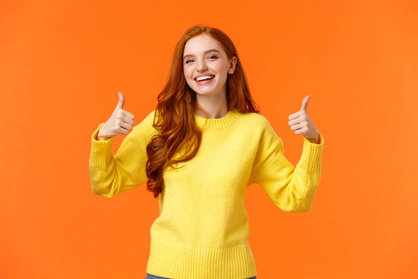 Holidays, gestures, people concept. Cheerful cute redhead girl smiling and showing thumbs-up recommend product, give positive reply, agree or like something, nod agreement, orange background - Photo, Image