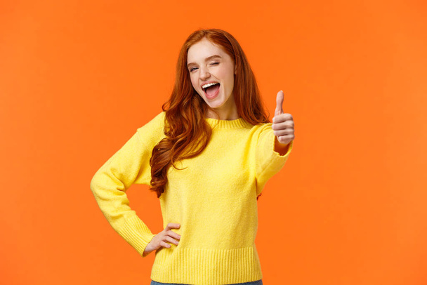 Diciendo que sí. Mujer pelirroja alegre en suéter amarillo dando gesto de pulgar hacia arriba y guiño, alentando a mantener, aprobando idea, expresar opinión positiva, recomendar producto o evento, fondo naranja
 - Foto, Imagen