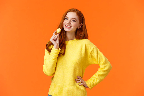 Талия портрет прекрасная рыжая девушка держит вкусный желтый макарон у рта и радостно улыбается, как едят сладости, купить десерт для романтического свидания валентинки, позируя веселый оранжевый фон
 - Фото, изображение