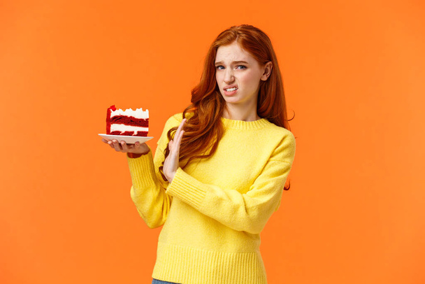 Walgelijk en ontevreden, kieskeurig roodharige meisje zorg voor lichaamsvorm, eet geen snoepjes, houdt stuk smakelijke taart vast en weigert te eten, toont stopbord, grimas op camera met afkeer, oranje achtergrond - Foto, afbeelding