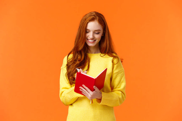 Pas-up portrét atraktivní zrzka dívka 20s psát do svého deníku, výplň-in-to-list nebo osobní rozvrh do nového notebooku, usmívat se, jak mají vše naplánované, stojící oranžové pozadí bezstarostné - Fotografie, Obrázek