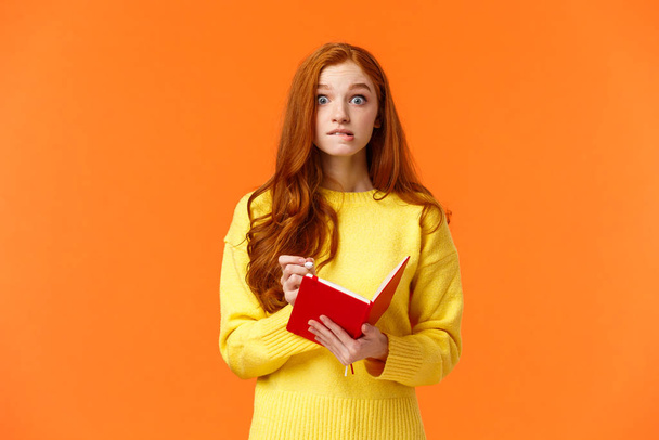 Troubled redhead caucasian girl forgot about appointment, remember task after looking notebook, biting lip and stare concerned or embarrassed, writing down important notes, orange wall - Photo, Image