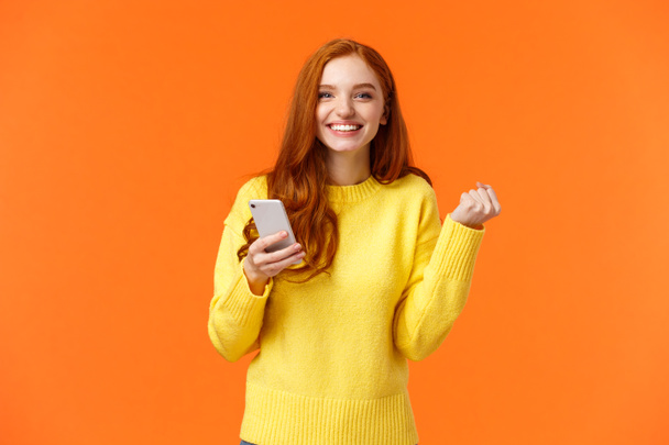 Happy girl share good news with you. Cheerful pretty redhead female holding smartphone, fist pump in triumph or celebration, winning in game, got message from guy she likes, orange background - Photo, Image