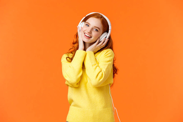 Portrait taille haute mignonne adolescente rousse en casque blanc, tête inclinée écouter de la musique, toucher les écouteurs et la caméra souriante, chanter le long de la chanson préférée, debout fond orange
 - Photo, image