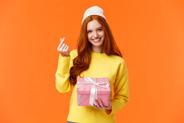 Lovely redhead girlfriend got gift from her girl, showing korean heart gesture and smiling silly, holding pink present box, wearing winter beanie, standing over orange background happy - Foto, afbeelding