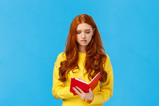 Perplexed and uneasy, concerned cute redhead female student having troubles with schedule, frowning reading girlfriend diary, feeling guilty, holding red notebook, feeling troubled, blue background - Photo, Image