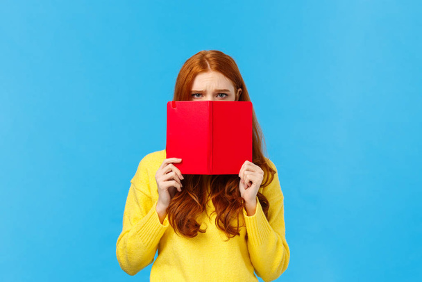 Upset gloomy and uneasy cute pouting redhead girl, frowning dont want speak to you, hiding face behind red notebook, frowning and staring offended, sulking over blue background, bad mood - Photo, Image