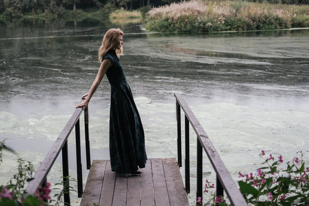 Портрет жінки, одягненої в голлівудське хвилясте волосся і довге плаття, стоячи на мосту біля красивого брудного ставка. - Фото, зображення