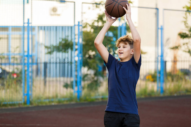 Cute boy in blue t shirt plays basketball on city playground. Active teen enjoying outdoor game with orange ball. Hobby, active lifestyle, sport for kids. - Photo, Image